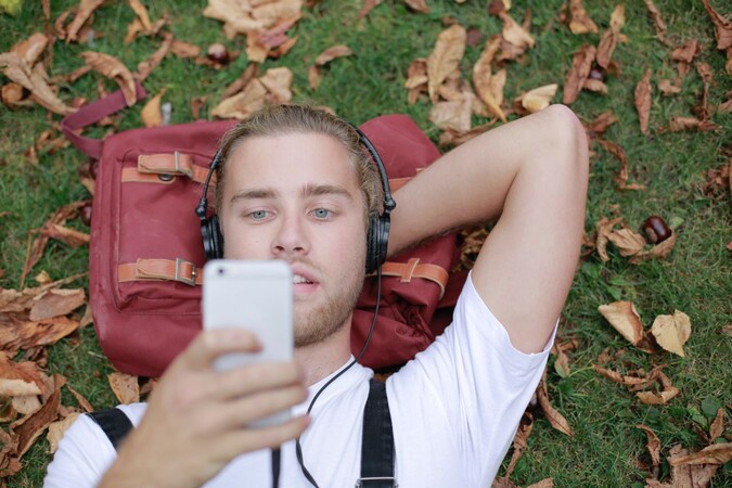 Ein junger Mann liegt auf dem Rasen. Er trägt Kopfhörer und schaut auf ein Smartphone. Umgeben ist er von herabgefallenen Laub, seinen Rucksack benutzt er als Kopfkissen.